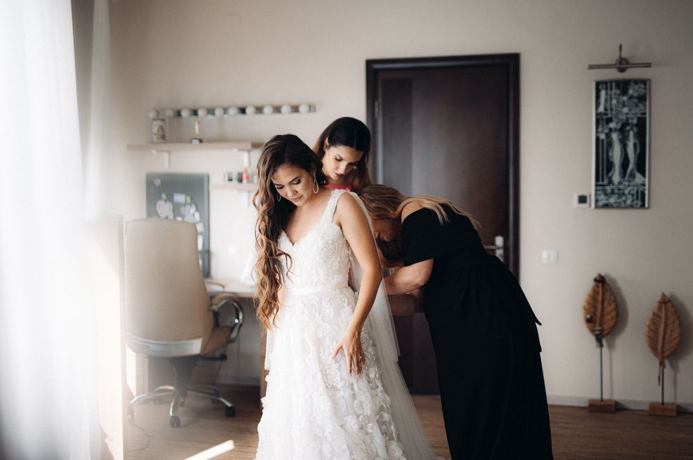 A candid moment of wedding dress fitting in a sunlit room with helpers making final adjustments.