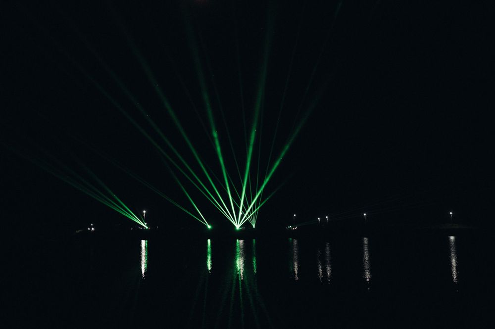 Dramatic green laser light beams reflect over dark water at night.