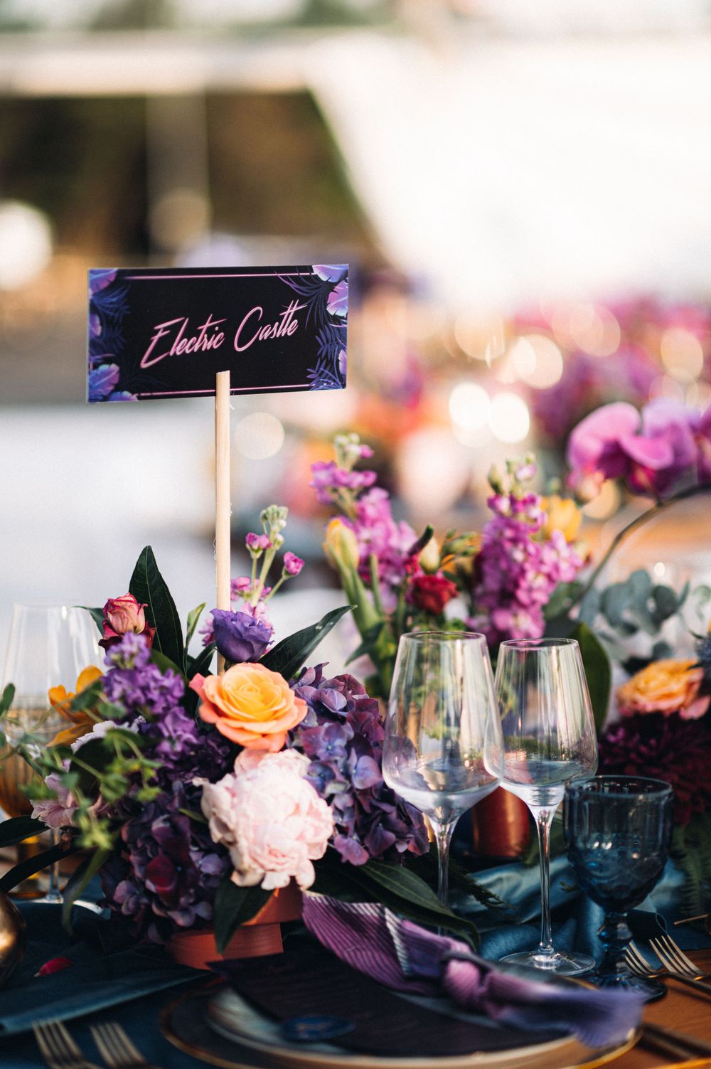 Close-up of wedding table decor featuring purple and orange flowers, wine glasses, and a decorative name card.