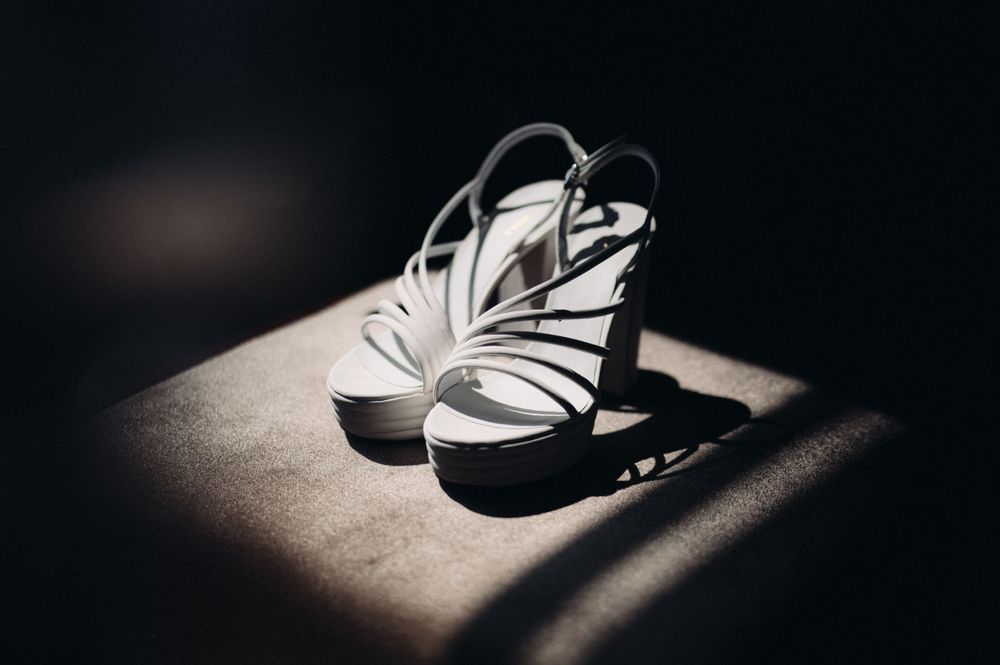 A dramatic black and white photo of white high heeled wedding shoes casting shadows on a textured surface.