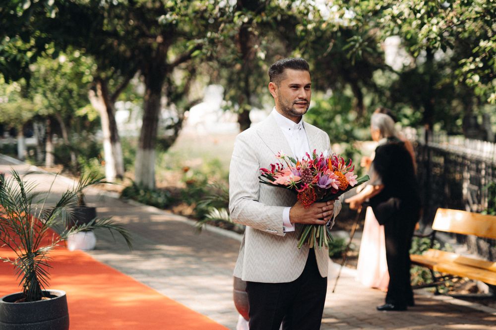 Groom in light blazer holding vibrant red and pink wedding bouquet on outdoor path.
