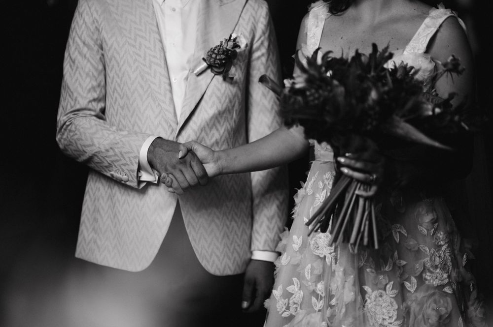 Black and white photo of newlyweds holding hands with a textured white jacket and lace dress.
