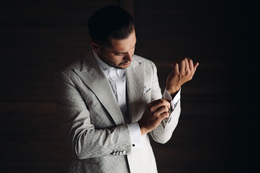 A dramatic photo of someone in a light suit adjusting their sleeves against a dark background.