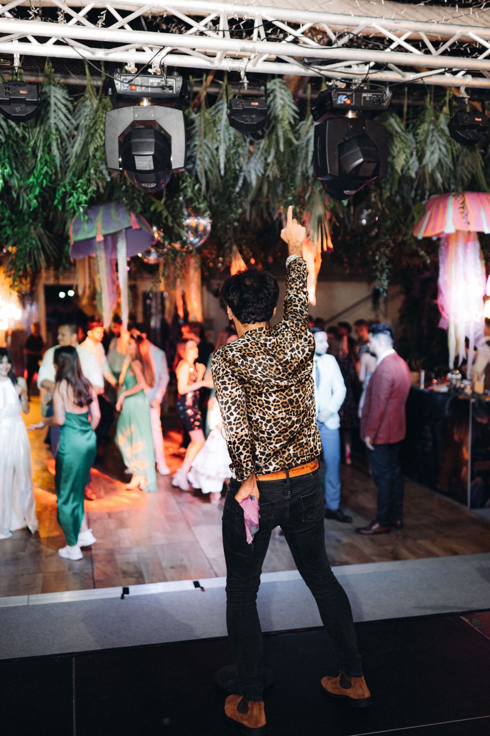 DJ performs at decorated venue with hanging lights and greenery above dance floor.