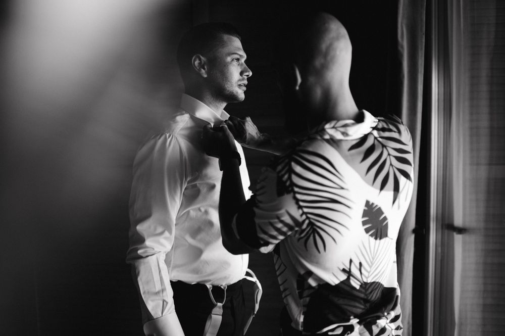 A moody black and white photo of two people in formal attire in intimate conversation by a window.