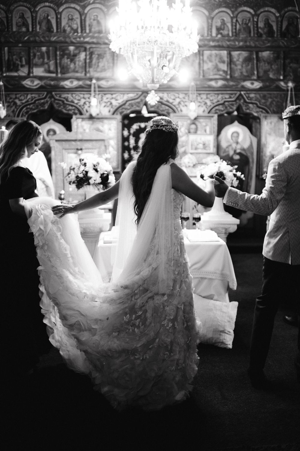 Black and white photo of bride in flowing lace gown during Orthodox church ceremony.