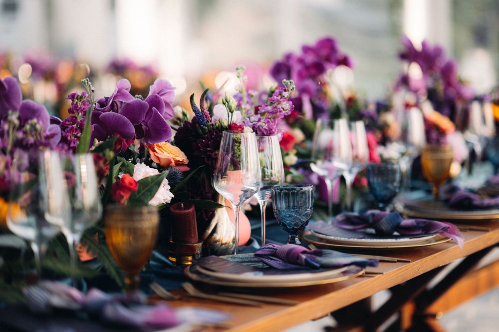 Detailed view of luxury place settings with purple florals and dark plates on wooden tables.