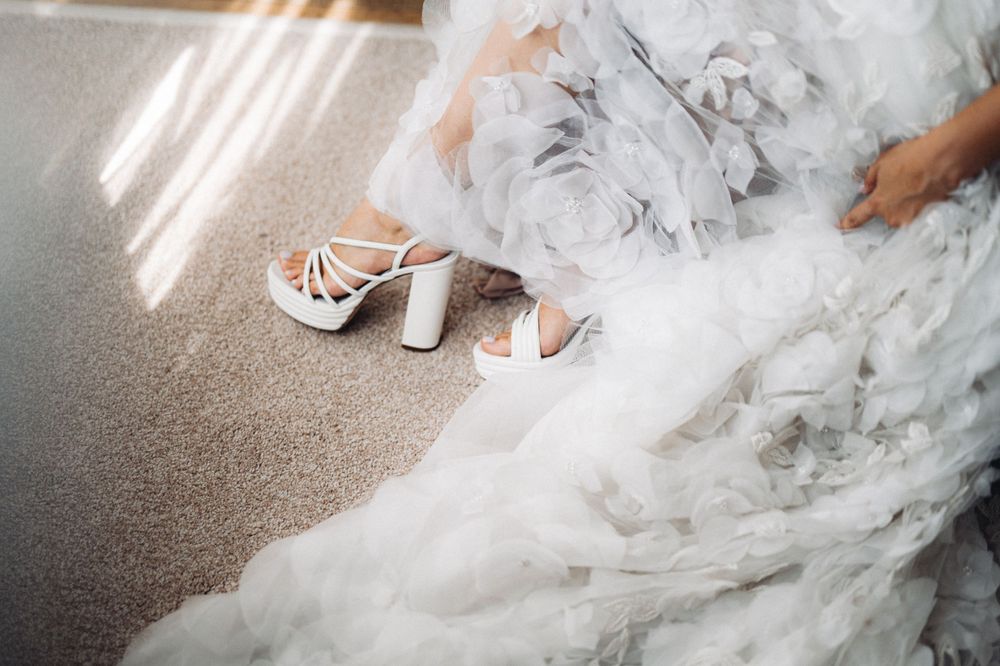 White strappy bridal heels rest beside the flowing tulle of a wedding dress on carpeted floor.