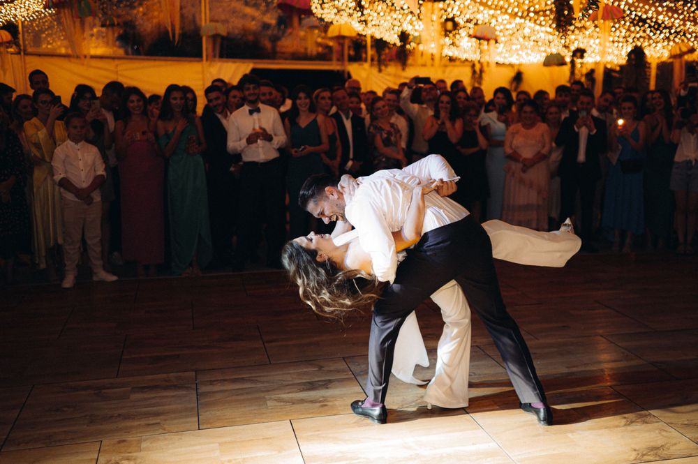 Elegantly dressed couple performs dramatic dip dance move on illuminated ballroom floor surrounded by guests.
