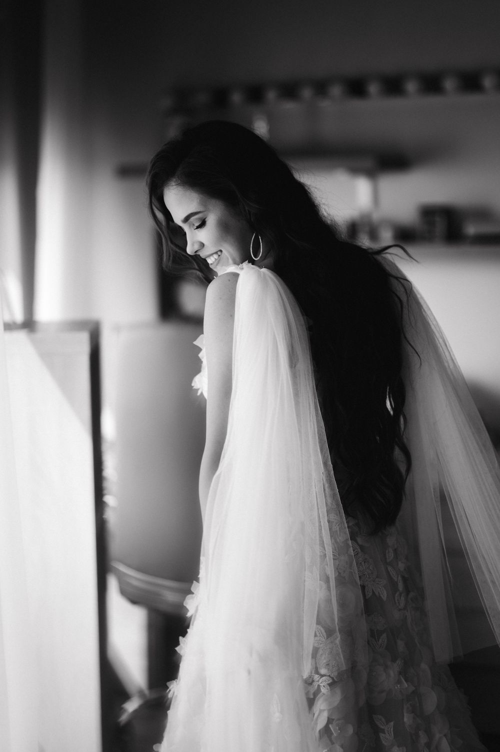 A black and white portrait of a bride with long dark hair looking down while wearing a lace gown and veil.