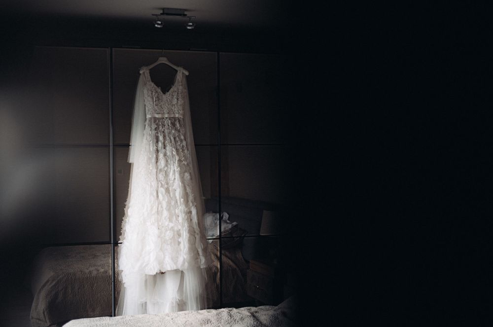 An elegant white lace wedding dress hanging in a dimly lit room.