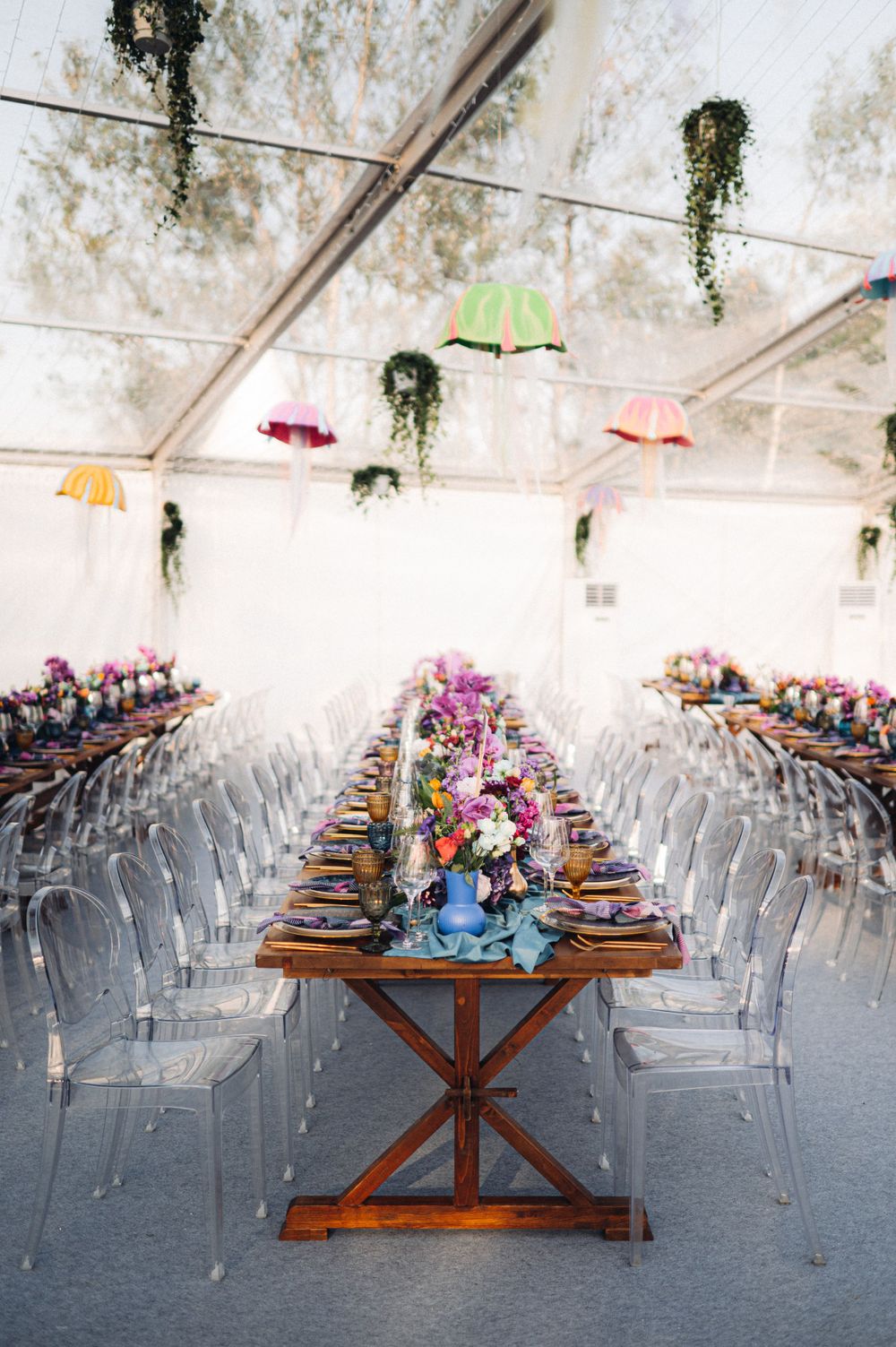 Modern wedding reception setup with long wooden tables, ghost chairs, and hanging greenery in a white tent.