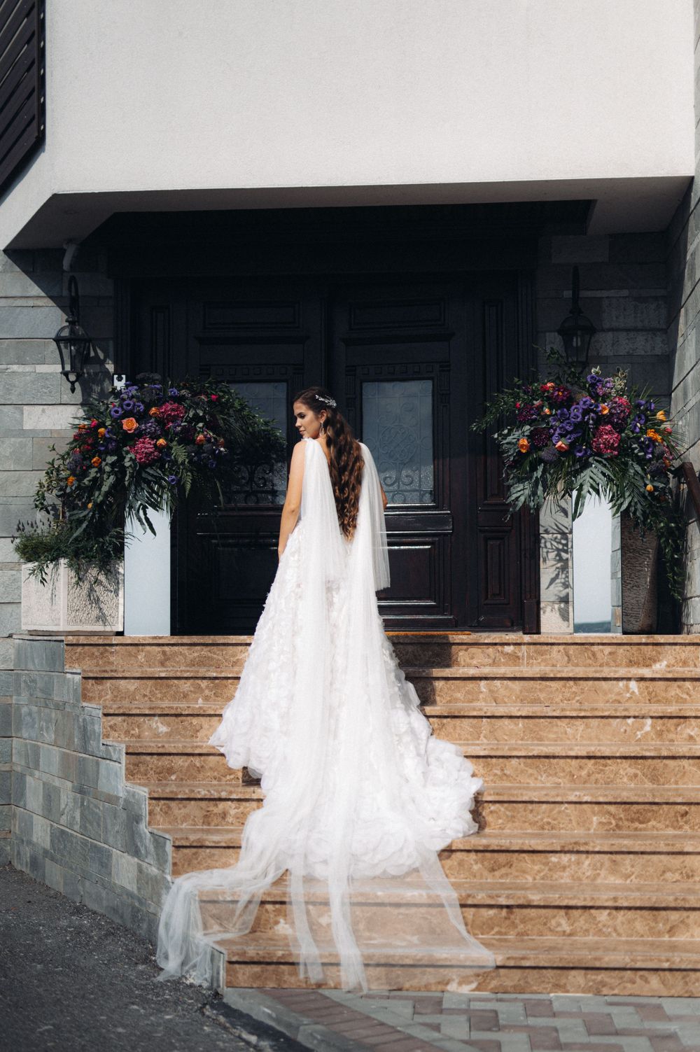 Bride in flowing white gown stands on stone steps with floral arrangements framing a black doorway.