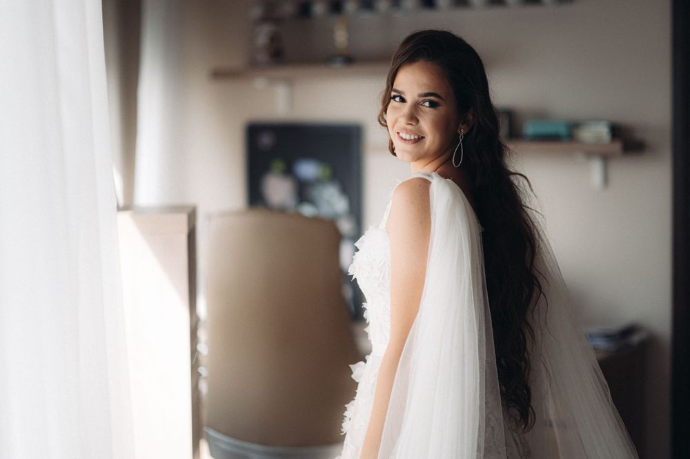 A bride in a white wedding dress with flowing veil smiles while looking over her shoulder near a window.