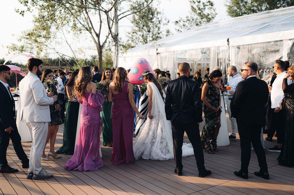Wedding guests mingle on an outdoor deck during sunset reception with white tent in background.