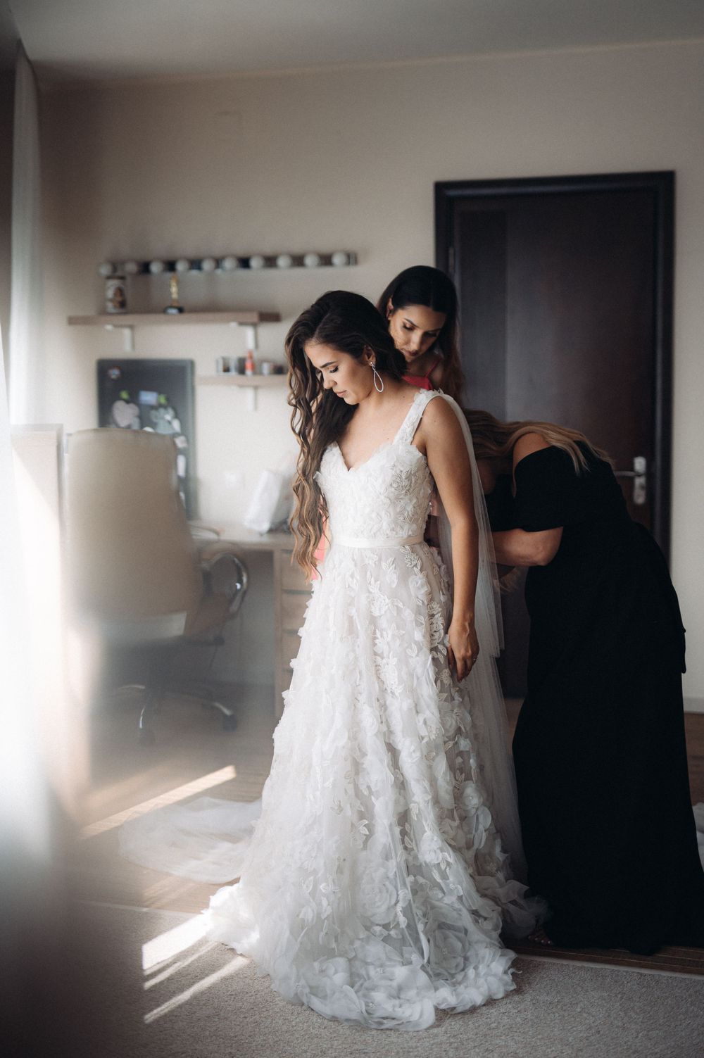 A serene bridal preparation scene with helpers adjusting a flowing lace wedding gown in natural light.