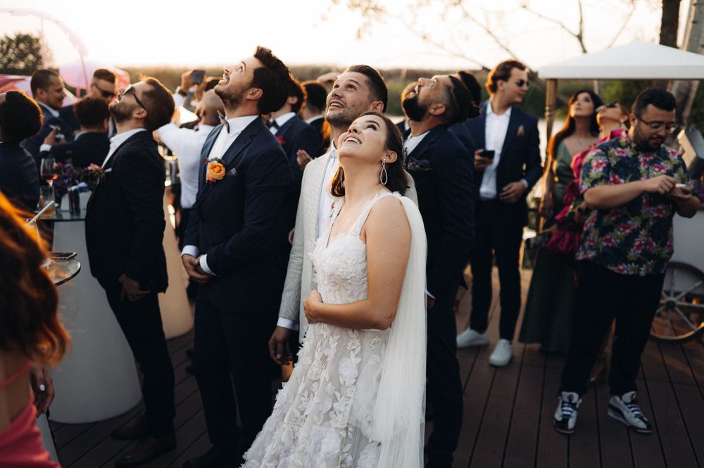 Outdoor wedding reception with guests gathered on wooden deck at sunset.