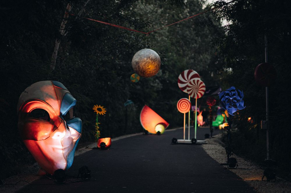 Illuminated art installation with glowing sculptures and pinwheels along a dark pathway.