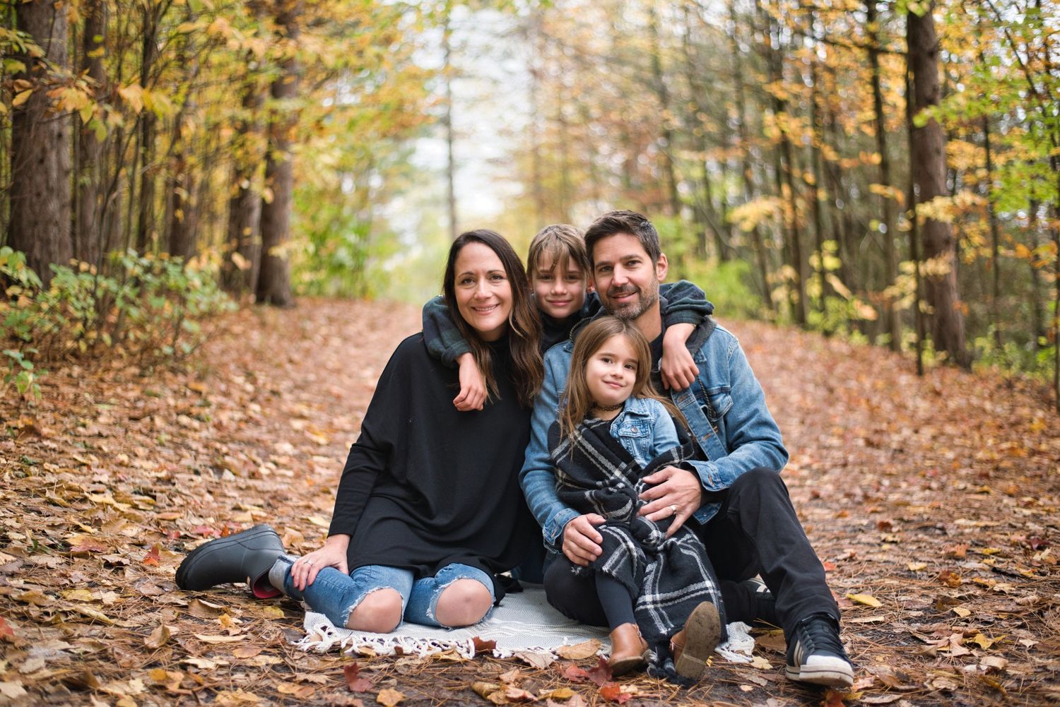 Family sitting together on fallen leaves during autumn forest photo session.