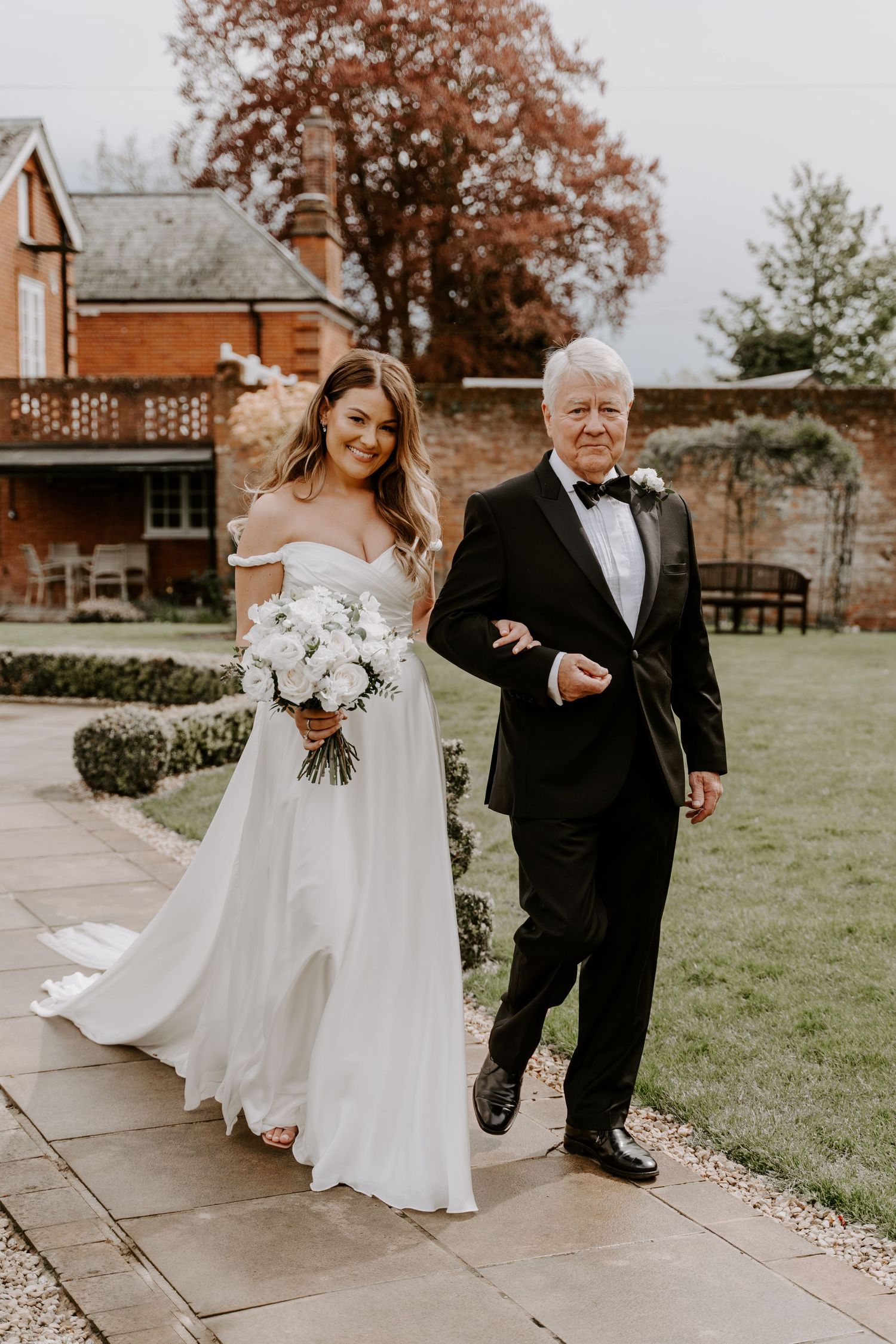 Bride walking through a venue garden linked arms with her dad. She is holding a white flower bouquet.