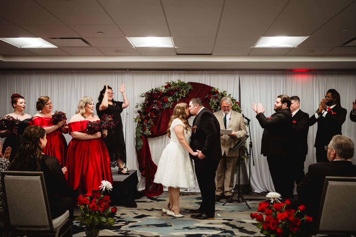 Wedding ceremony moment captured in indoor venue with red-dressed attendants and white decor.