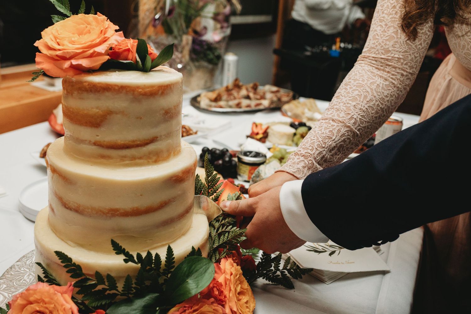 Semi-naked wedding cake decorated with orange roses and greenery being cut.