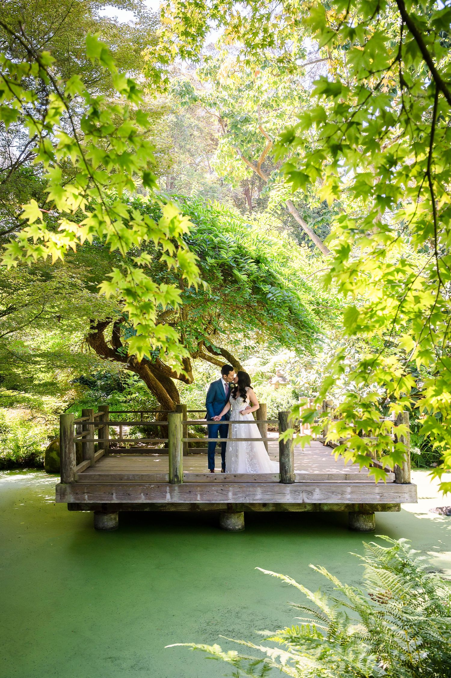 A couple in wedding attire stands on a wooden deck overlooking serene green waters.