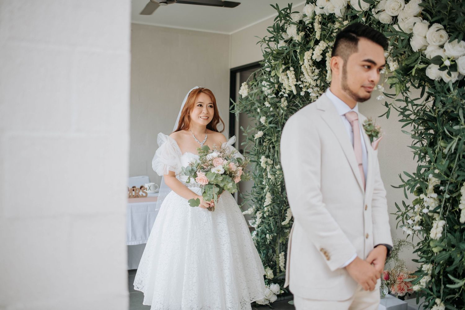 Wedding couple stands before floral arch during ceremony preparation.