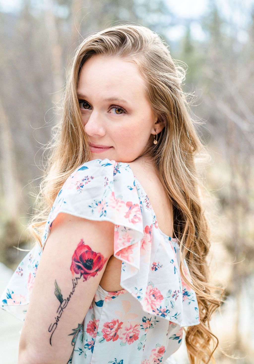 Woman in floral dress showing rose tattoo on arm with blurred nature background.