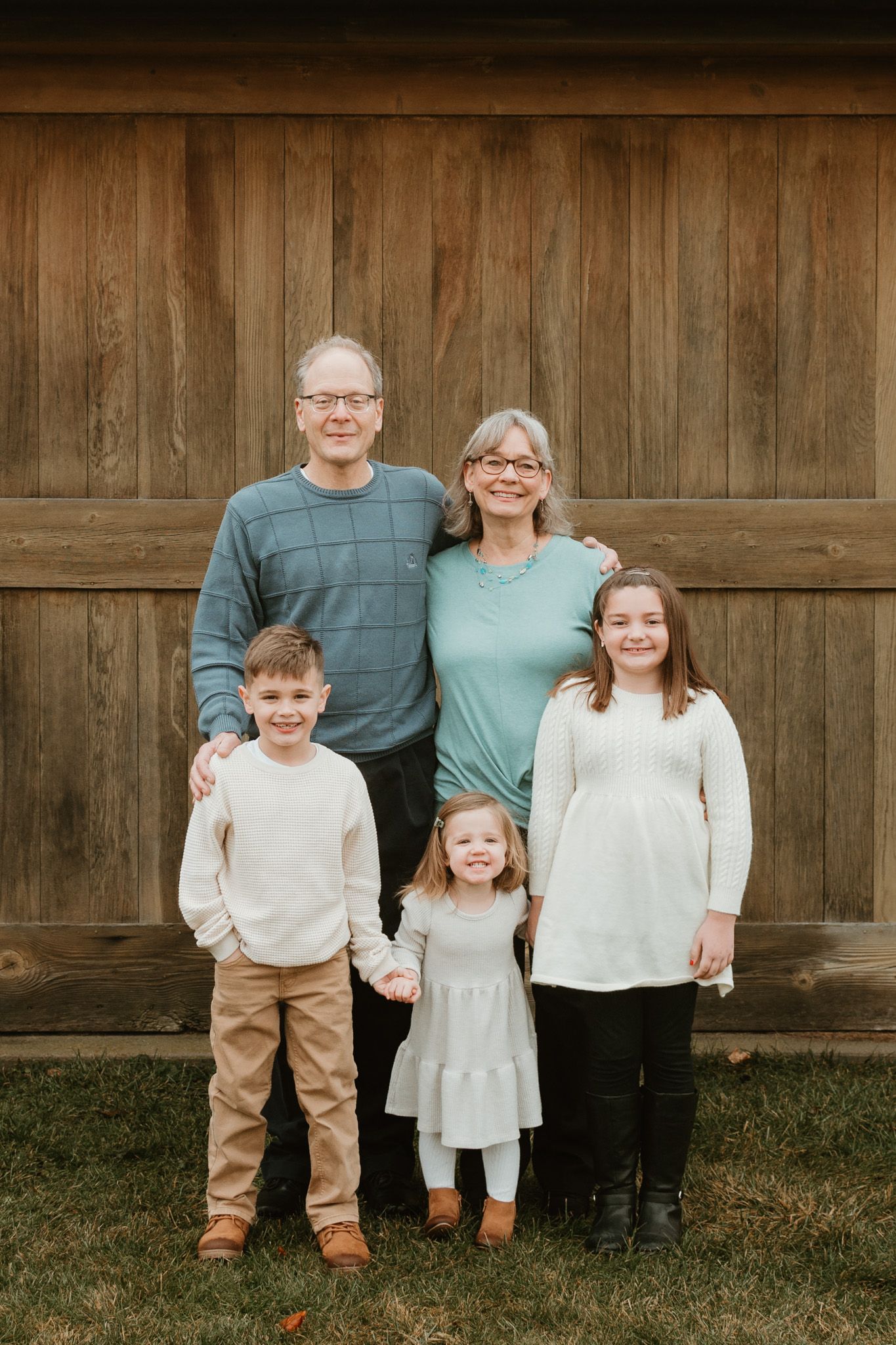 Grandparents pose with three grandchildren in front of a rustic wooden barn backdrop outdoors.