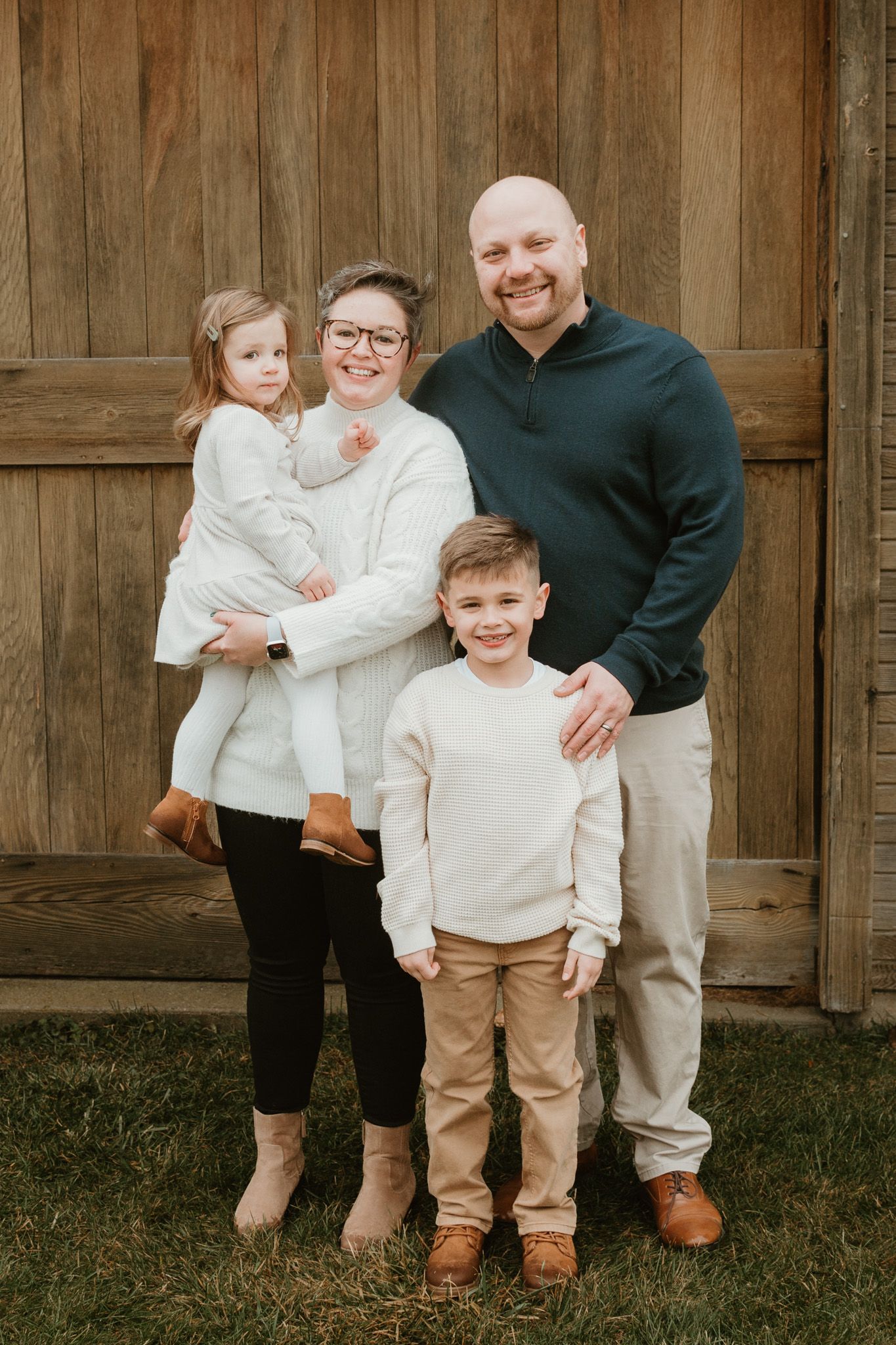 Happy family of four with two young children smiling together in front of a rustic wood wall.