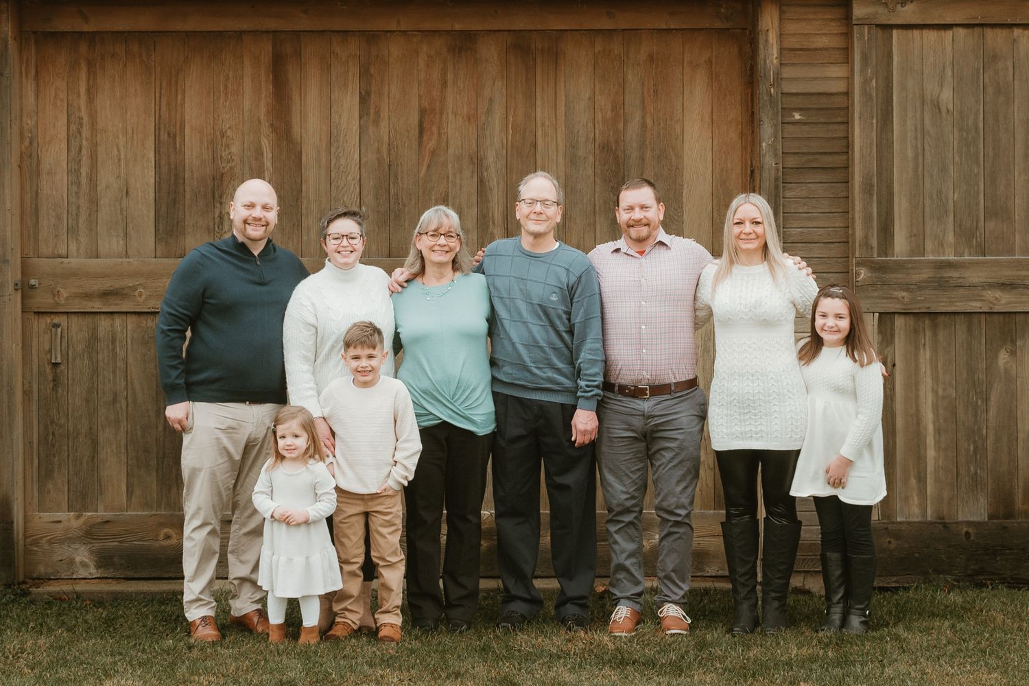 Large family portrait of eight people smiling in front of rustic wooden barn wall outdoors.