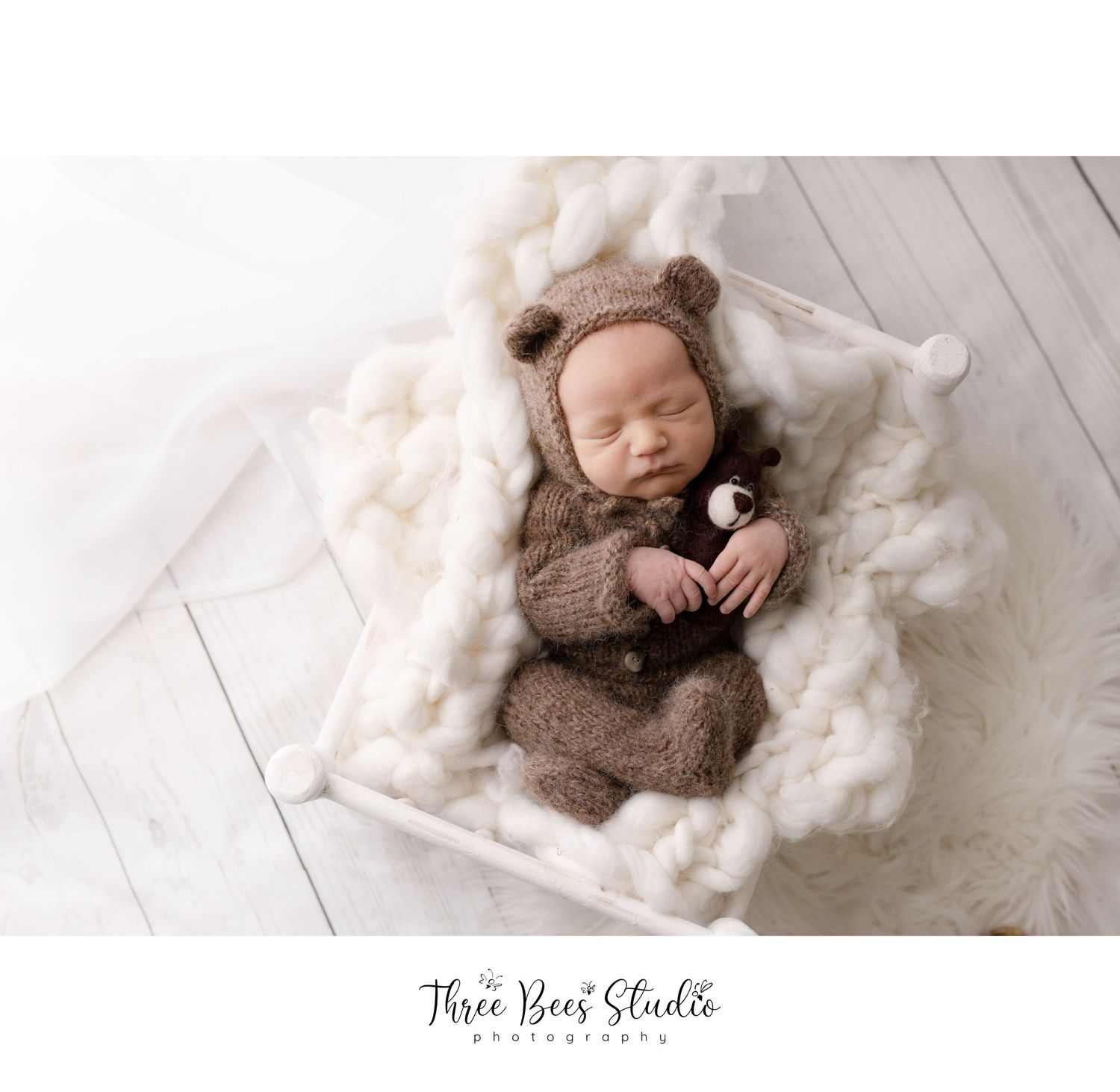 Newborn baby wearing brown teddy bear outfit sleeping on white blanket.