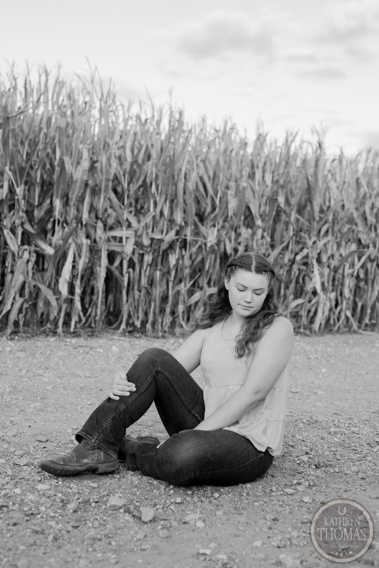 Black and white portrait of someone sitting by a corn field on a dirt path wearing dark clothes and boots.