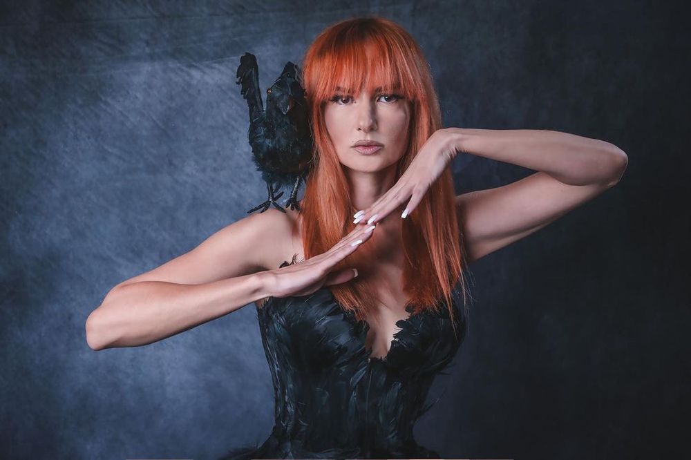A woman with red hair poses elegantly in a black feathered outfit, with a black raven perched on her shoulder against a dark backdrop