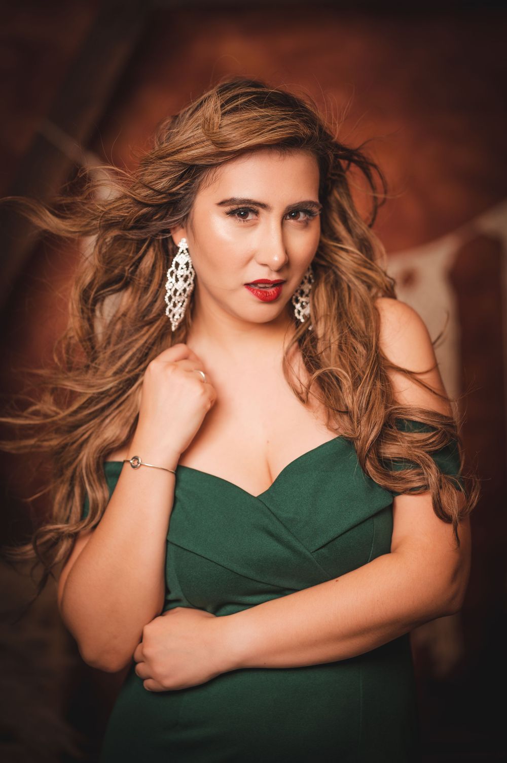A woman in a deep green off-shoulder dress poses with windblown hair, bold red lips, and statement earrings against a warm-toned background.