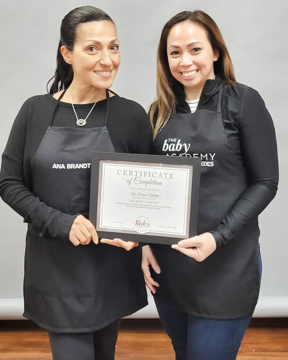 Two photographers wearing black aprons display a certificate in a studio setting.