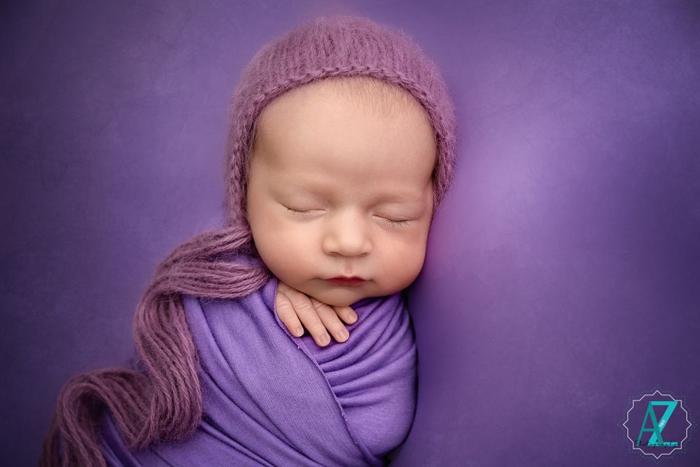 Peaceful newborn wearing knit hat swaddled in purple fabric against purple backdrop.