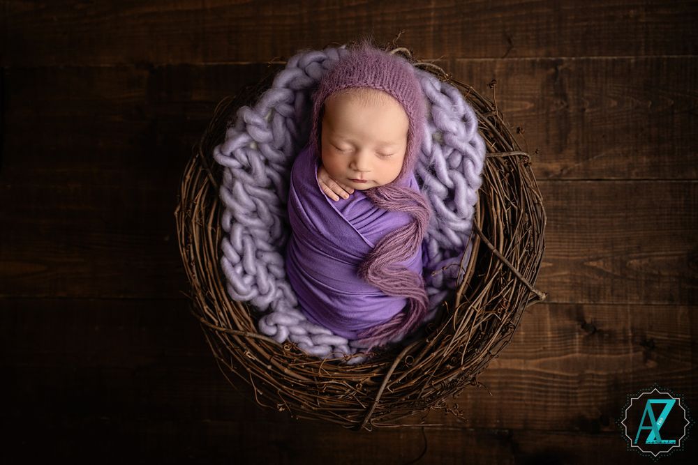 Newborn baby swaddled in purple fabric sleeping peacefully in a rustic wooden nest with cozy knit blanket.