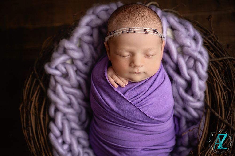 Sleeping newborn wrapped in purple swaddle wearing delicate headband in wooden nest with knit blanket.