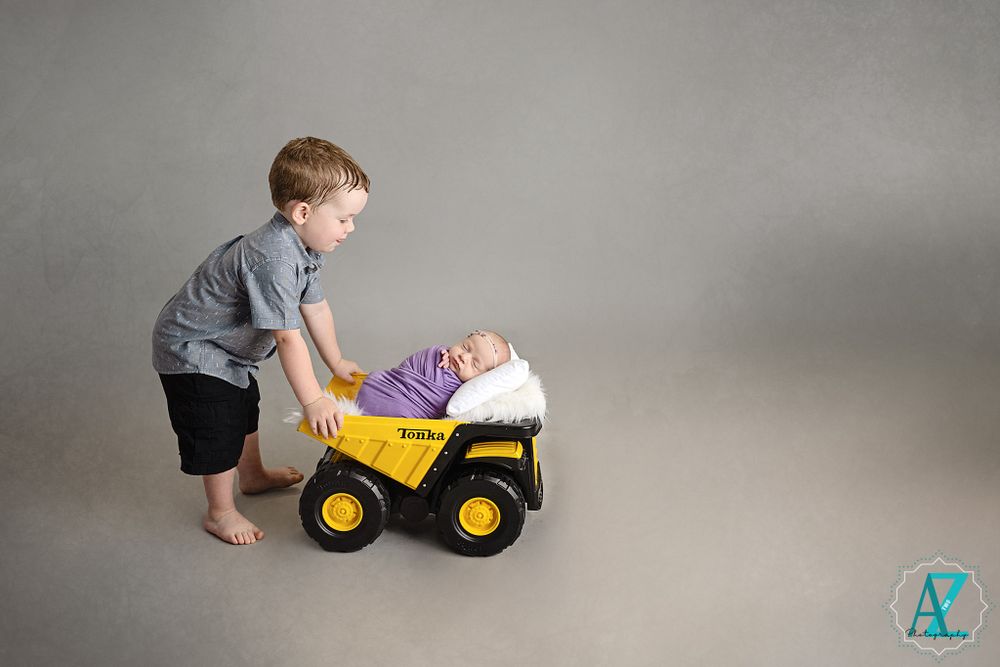 Young child pushing toy dump truck with baby sibling inside against grey backdrop.
