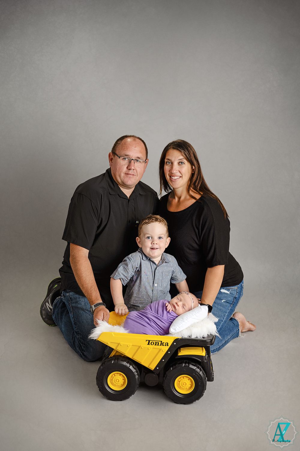 Family portrait with parents and child posing with yellow toy dump truck against grey backdrop.