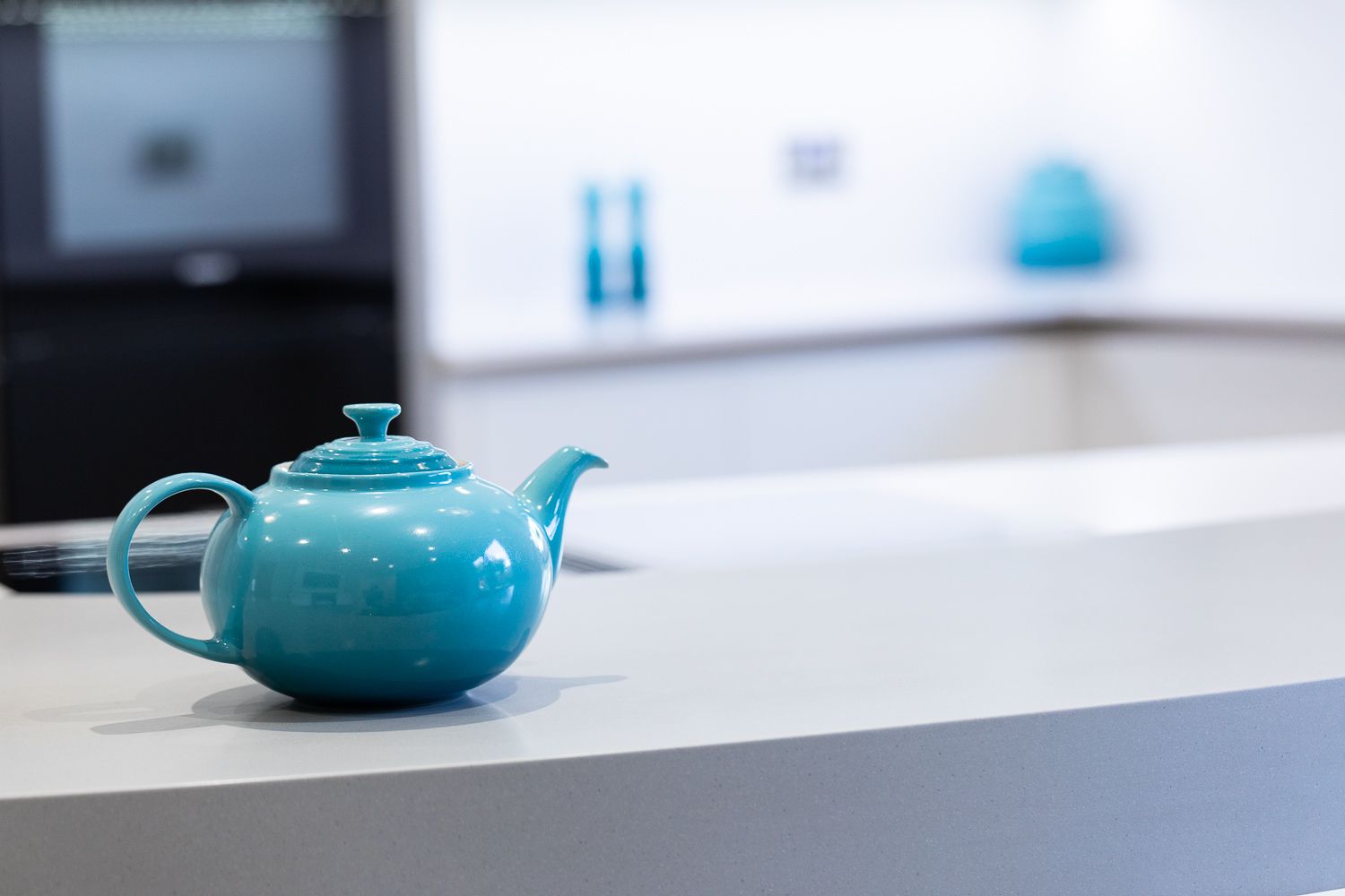 A teal ceramic teapot sits on a gray countertop with blurred kitchen decor in the background.