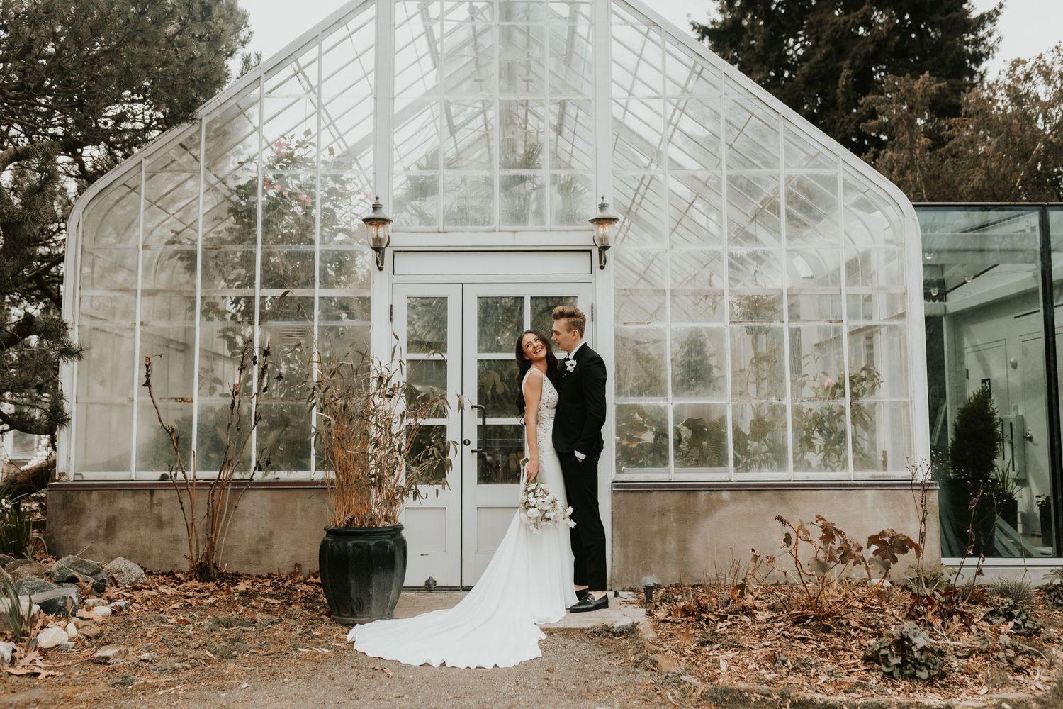A couple shares an intimate moment in front of a glass greenhouse.