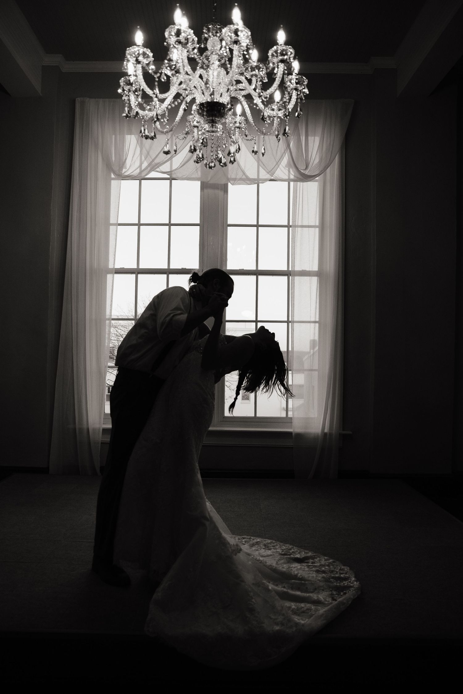 Silhouetted couple embracing under crystal chandelier by window in dramatic wedding photo.