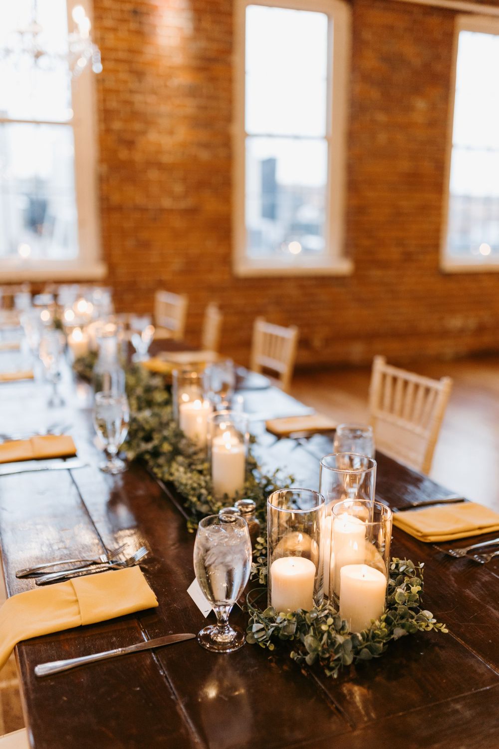 Rustic wedding head table with white candles, greenery runner, and gold napkins in brick venue.