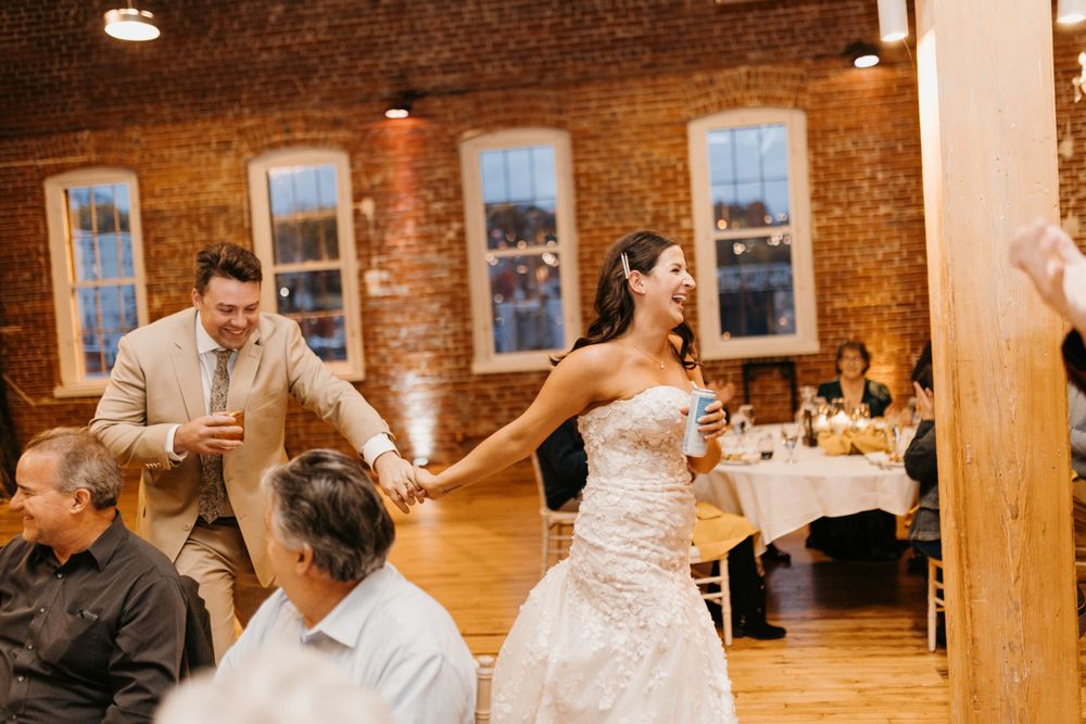 Bride and groom enter wedding reception hall with brick walls, smiling and holding hands.
