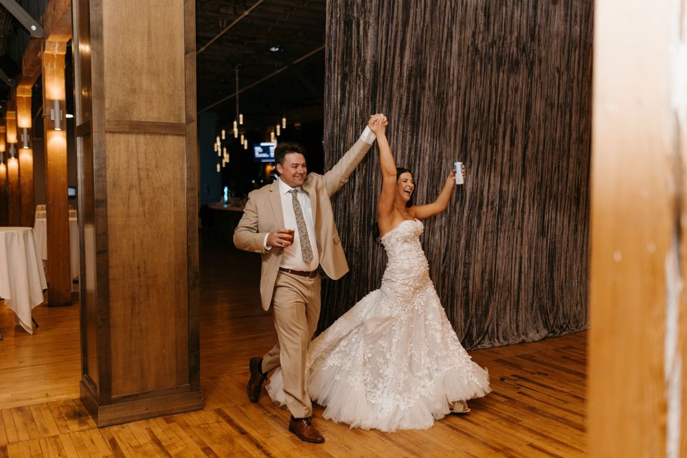 Bride and groom make grand entrance into wedding reception raising hands and holding drinks joyfully.