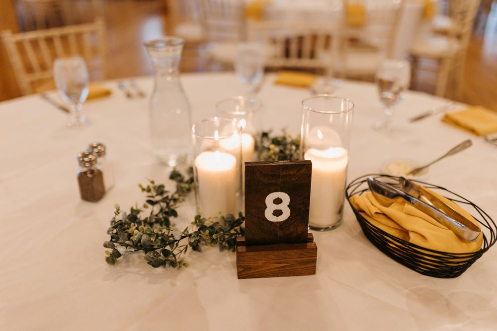 Wedding table centerpiece with white candles, greenery, and wooden table number 8 on white linen.