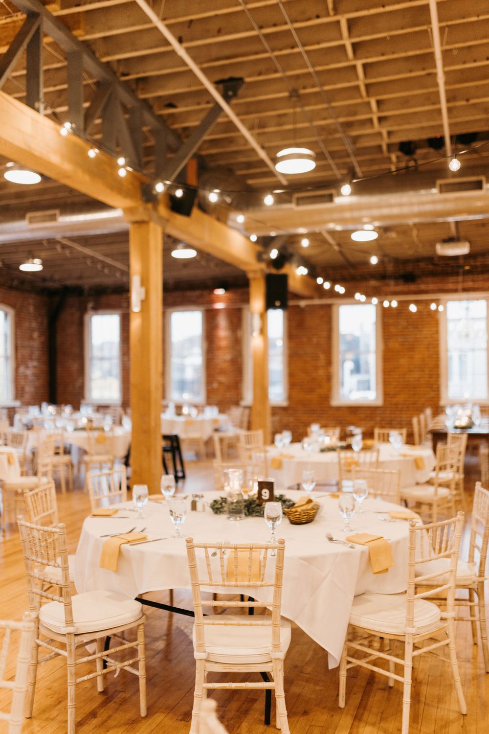 Elegant wedding reception hall with white round tables, chiavari chairs, string lights, and brick walls.